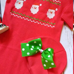 May include: Red baby onesie and matching hat set. The onesie features an embroidered design of three Santa faces with red hats and white beards. The hat has a green bow with white polka dots. A festive holiday outfit.
