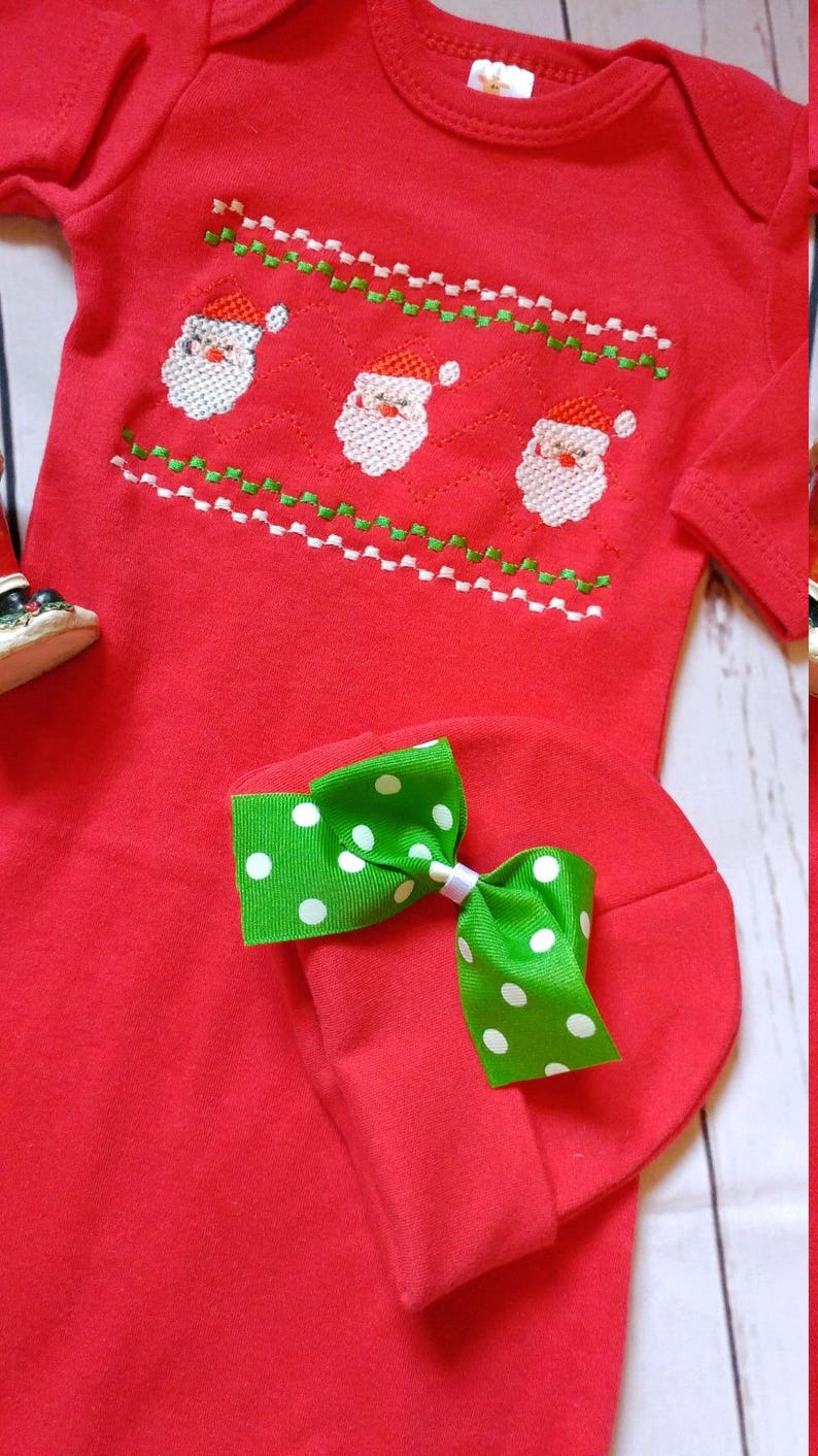 May include: Red baby onesie and matching hat set. The onesie features an embroidered design of three Santa faces with red hats and white beards. The hat has a green bow with white polka dots. A festive holiday outfit.