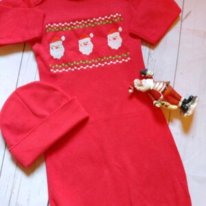 May include: Red baby sleeper and matching cap. The sleeper features an embroidered design of three Santa faces with white beards and red hats. A small Santa figurine is also present.