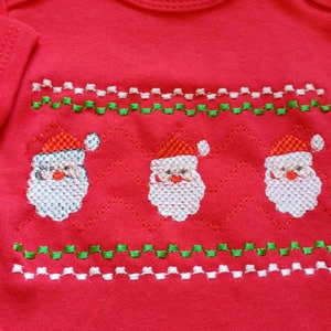 May include: Red baby bodysuit with an embroidered Santa Claus design. The design features three Santas with white beards and red hats, along with green and white zigzag patterns. A festive garment for the holiday season.
