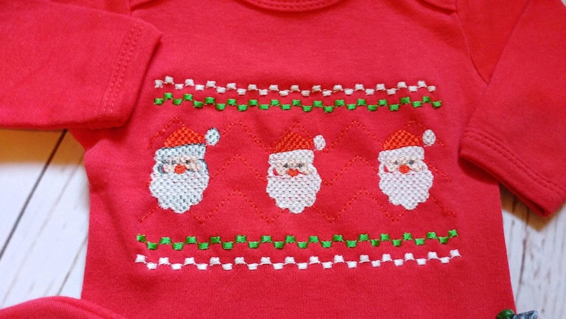May include: Red baby bodysuit with an embroidered Santa Claus design. The design features three Santas with white beards and red hats, along with green and white zigzag patterns. A festive garment for the holiday season.