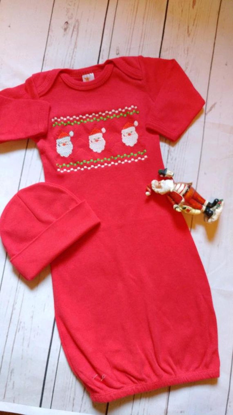 May include: Red baby Christmas outfit with a matching hat. The outfit features an embroidered design of three Santa faces with white beards and red hats. A small Santa figurine is next to the outfit.
