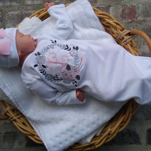May include: A white baby gown with long sleeves and a pink and gray floral embroidery design featuring a letter "L". The gown is worn by a baby wearing a pink and white hat with a bow.