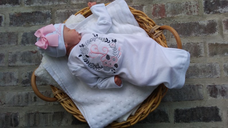 May include: A white baby gown with long sleeves and a pink and gray floral embroidery design featuring a letter "L". The gown is worn by a baby wearing a pink and white hat with a bow.
