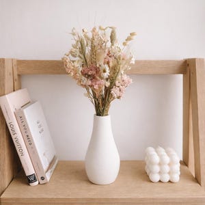 May include: A white ceramic vase displays a bouquet of dried flowers in pink, beige, and green hues. The vase is on a light wood shelf with two books and a bubble cube candle. The books are titled "LOUIS VUITTON" and "ALL YOU NEED IS LOVE".