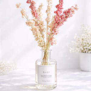 May include: A clear glass diffuser bottle labelled "DAISY" containing dried flowers. The flowers are in shades of pink and cream, arranged vertically. The overall aesthetic is delicate and natural, suitable for home decor.