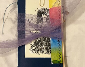 May include: A dark blue journal with a purple tulle ribbon tied around it. The journal has a paperclip holding a page with a black and white illustration of a woman with wings. The journal has a small crocheted flower on the cover.