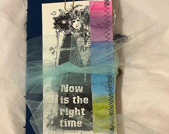 May include: A dark blue journal with a crocheted flower bookmark. The cover is decorated with a black and white image of a woman with a crown of flowers, a blue and green watercolor background, and the text "Now is the right time".
