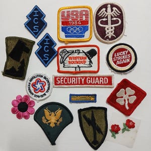 May include: A collection of vintage embroidered patches, including a red, white, and blue USA patch with the 1984 Olympics logo, a green patch with a four-leaf clover, a red patch with a shamrock, a red, white, and blue patch with the text "Western Roundup", a red patch with the text "Lucky Strikes Again", a blue patch with the text "ICS", a red, white, and blue patch with the text "American Revolution Bicentennial 1776-1976", a red and white patch with the text "Security Guard", a green patch with a golden eagle, a green patch with a plane, and a brown patch with a silhouette of a dog.
