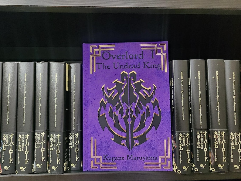 Pode incluir: Um livro encadernado em roxo com letras douradas intitulado "Overlord I The Undead King" de Kugane Maruyama. O livro est&aacute; numa estante com outros livros.