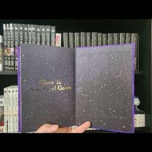 Pode incluir: Um livro encadernado em roxo com uma capa preta e letras douradas que dizem "Glory To Ainz Ooal Gown". A capa tem um design de noite estrelada.