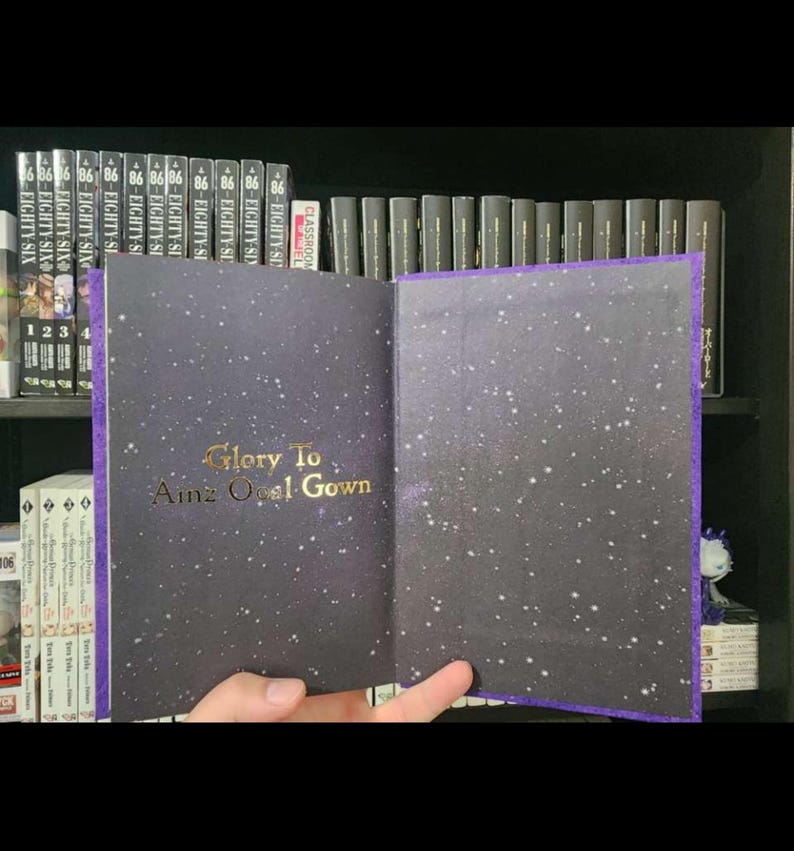 Pode incluir: Um livro encadernado em roxo com uma capa preta e letras douradas que dizem "Glory To Ainz Ooal Gown". A capa tem um design de noite estrelada.