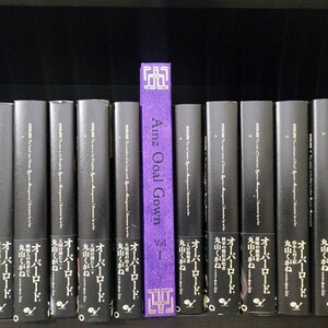 Pode incluir: Uma estante com uma fila de livros pretos com texto branco. O livro no centro &eacute; roxo com texto dourado que diz "Ainz Ooal Gown Vol 1".