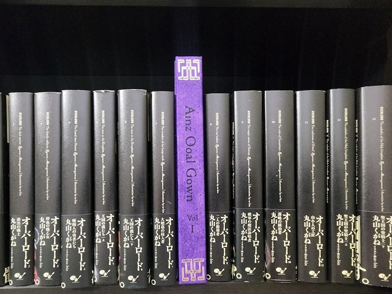 Pode incluir: Uma estante com uma fila de livros pretos com texto branco. O livro no centro &eacute; roxo com texto dourado que diz "Ainz Ooal Gown Vol 1".