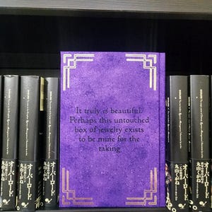 Pode incluir: Um livro roxo com detalhes dourados e o texto "It truly is beautiful. Perhaps this untouched box of jewelry exists to be mine for the taking." O livro est&aacute; em uma estante com outros livros.
