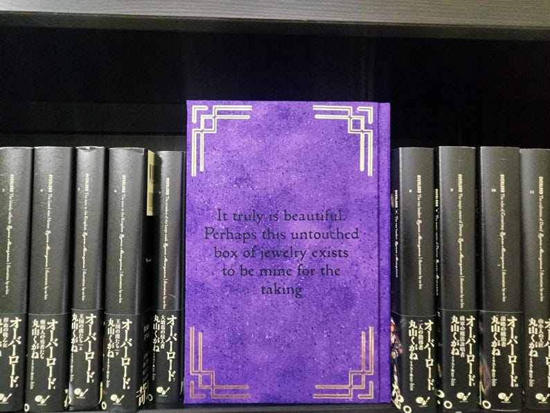 Pode incluir: Um livro roxo com detalhes dourados e o texto "It truly is beautiful. Perhaps this untouched box of jewelry exists to be mine for the taking." O livro est&aacute; em uma estante com outros livros.