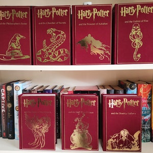 May include: A bookshelf with a collection of red hardcover books with gold lettering. The books are all Harry Potter novels, with titles like "Harry Potter and the Sorcerer's Stone" and "Harry Potter and the Chamber of Secrets".