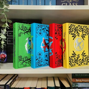 Puede incluir: Cuatro libros de bolsillo con diseños florales en blanco y negro en las portadas. Los libros se titulan "Mockingjay", "The Hunger Games" y "Catching Fire". Los libros están dispuestos en una fila en una estantería.