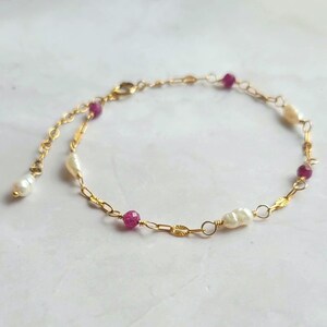 May include: A delicate gold chain bracelet with alternating freshwater pearls and faceted ruby beads. The bracelet has a lobster clasp and a small pearl drop. The gold chain is linked with small, flat, oval-shaped links.