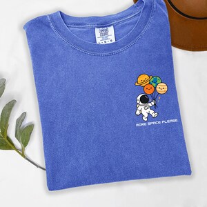 May include: A blue short-sleeve t-shirt with a graphic on the chest featuring a white astronaut holding a cluster of four colorful planet balloons with smiling faces. Below the graphic, the text reads MORE SPACE PLEASE in white capital letters.