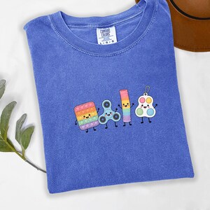 May include: A blue short-sleeved t-shirt with a Comfort Colors label at the neck. The centre features an embroidered graphic of four colourful, smiling fidget toys: a square pop-it, a blue spinner, a rainbow spring, and a white circular pop-it.