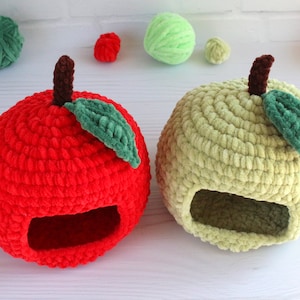 Op de afbeelding: Twee gehaakte appelvormige huisdierbedden, een rood en een groen, met een kleine opening om binnen te komen. De bedden zijn gemaakt van zacht, pluche garen.