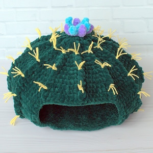 May include: A green crocheted cactus-shaped pet bed with yellow yarn accents and a purple and blue flower on top.