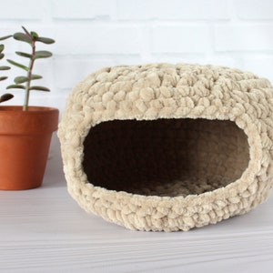 Puede incluir: Una cama para mascotas de crochet beige de forma redonda con un agujero en la parte delantera para la entrada. La cama está hecha de un material suave y esponjoso.