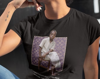 May include: A black t-shirt featuring a graphic of a person sitting on a chair against a purple patterned square background. Below the image, the text reads: I cant believe what you say, because I see what you do - James. The shirt has short sleeves.
