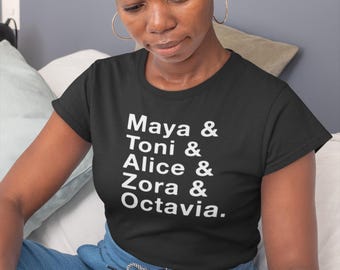 May include: Black t-shirt with white text listing names: Maya & Toni & Alice & Zora & Octavia. The person in the image is wearing a leopard print headband and gold hoop earrings. The person is reading a book and wearing blue pants.
