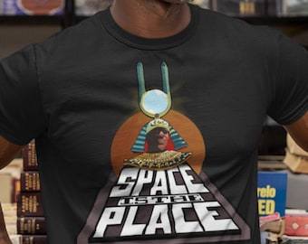 May include: A black short-sleeved t-shirt featuring a graphic print of a figure wearing an Egyptian-style headdress and collar. Below the figure, a large triangle contains the white text "SPACE IS THE PLACE" set against a dark orange circle.