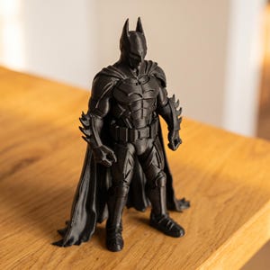 Könnte beinhalten: Eine mattschwarze, 3D-gedruckte Superhelden-Figur mit spitzer Maske, Brustemblem, Gürtel und einem langen, fließenden Umhang. Die Figur steht in einer starren Pose und weist detaillierte Rüstungselemente an Armen, Beinen und am Oberkörper auf.