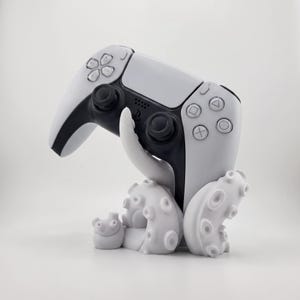 Könnte beinhalten: Ein weiß-schwarzer Videospiel-Controller, der von einem matten, weißen Krakenarm-Ständer gehalten wird. Der Controller verfügt über standardmäßige Richtungstasten, zwei Analogsticks und das PlayStation-Logo in der Mitte.