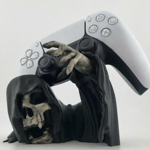 Könnte beinhalten: Eine schwarz-graue Sensenmann-Figur mit skelettiertem Gesicht und Kapuzenumhang, die als Controller-Ständer dient. Die Figur zeigt eine skelettierte Hand, die nach oben greift, um einen weißen und schwarzen kabellosen Game-Controller zu halten.
