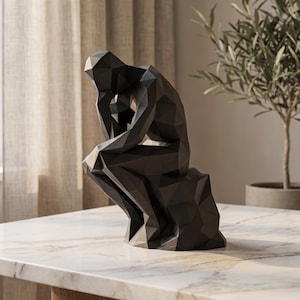 May include: A matte black sculpture of a seated figure in a contemplative pose, featuring a geometric, faceted design. The figure is depicted with its chin resting on one hand, with sharp angular planes forming the body and the base it sits upon.