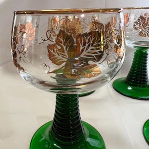 Puede incluir: Copas de vino transparentes con dise&ntilde;os de hojas de oro y vides. Las copas tienen una base y un tallo verdes. El borde de la copa est&aacute; adornado con oro. El tallo tiene un dise&ntilde;o acanalado.