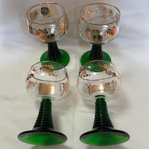 Puede incluir: Cuatro copas de vino transparentes con bordes dorados y dise&ntilde;os de racimos de uvas. Los tallos son negros con bases verdes. Las copas est&aacute;n dispuestas en dos filas de dos.