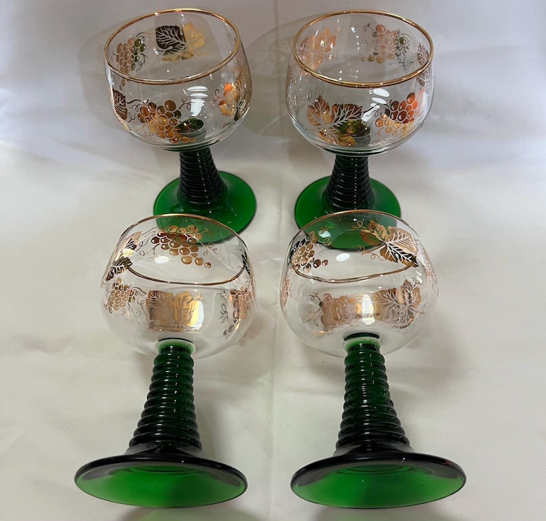 Puede incluir: Cuatro copas de vino transparentes con bordes dorados y dise&ntilde;os de racimos de uvas. Los tallos son negros con bases verdes. Las copas est&aacute;n dispuestas en dos filas de dos.