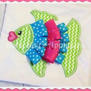 May include: Green and white chevron fish applique with a blue and white polka dot ribbon bow and a pink ribbon bow. The applique is on a white t-shirt. "Baby Kay's Appliques" is written in black text on the shirt.