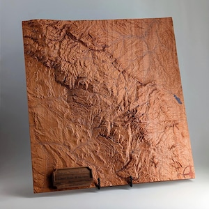 Könnte beinhalten: Eine dreidimensionale Reliefkarte des Flathead National Forest und der Great Bear Wilderness. Die Karte hat eine warme braune Farbe mit detaillierten topografischen Merkmalen. Eine kleine Holzplakette mit Text ist angebracht. Die Karte wird auf einem schwarzen Ständer präsentiert.