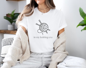 Op de afbeelding: Wit T-shirt met een zwart-witte illustratie van een bol garen en breinaalden. De tekst "in my knitting era" is onder de illustratie gedrukt.
