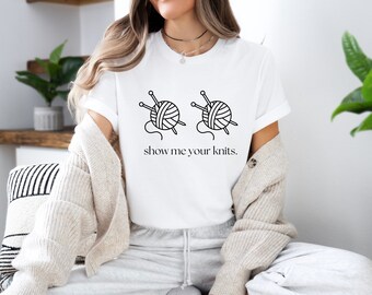 May include: White t-shirt with a black graphic of two yarn balls with knitting needles and the text "show me your knits." The shirt is worn by a person wearing a beige cardigan and gray sweatpants.