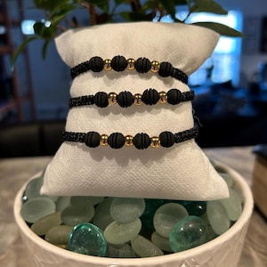 May include: Three black braided bracelets with gold beads. The bracelets are stacked on top of each other and are sitting on a white surface.