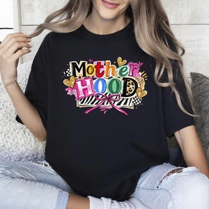 May include: Black t-shirt with the word "Motherhood" in colorful, playful lettering. The design includes hearts, a zebra print, and a pink bow. The shirt is worn by a person.