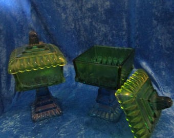 Puede incluir: Tres bomboneras de cristal verde con tapas. Los platos son cuadrados con un borde festoneado y se asientan sobre una base de pedestal. Las tapas tienen un pequeño asa en la parte superior. Los platos están dispuestos sobre un fondo azul.