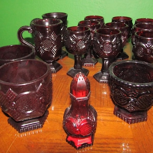 May include: A collection of ruby red glass cups, bowls, and a salt shaker with a textured, embossed design. The items are arranged on a wooden surface.