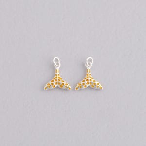 Könnte beinhalten: Zwei silberfarbene Walflossen-Anhänger mit gelben und orangefarbenen Punktmustern. Jeder Anhänger hat oben eine kleine Schlaufe zum Befestigen an Schmuck. Die Anhänger sind vor einem hellgrauen Hintergrund platziert.