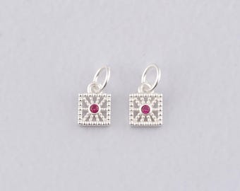 Puede incluir: Dos colgantes cuadrados plateados con un diseño de filigrana y una gema roja central. Cada colgante tiene un pequeño lazo en la parte superior para sujetarlo a una pulsera o collar. Los colgantes se muestran sobre un fondo gris claro.