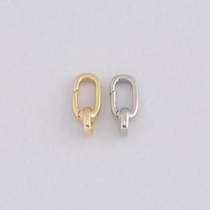 May include: Two metal oval-shaped clasps. One is gold-colored, the other is silver-colored. Each clasp has a small, rounded loop attached to the bottom. The clasps are likely for jewelry making or other craft projects.