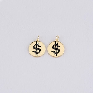 May include: Two gold-colored round pendants, each featuring a black dollar sign symbol. The pendants have a small loop at the top for attaching to a chain or other jewelry component. The background is a plain white.